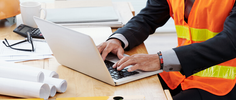 Homem de terno e colete sinalizador utilizando computador em obra.